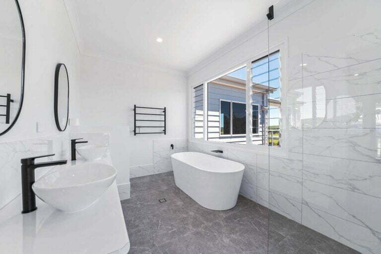A modern master bathroom with neutral floor to wall tiles, floor standing bath, his and hers vanity, black fixtures and a walk in shower.