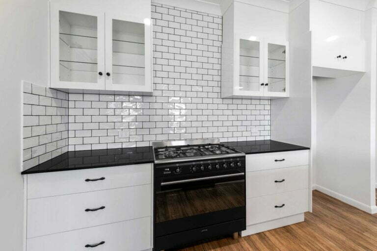 Large black Gas Cooktop and Oven with stainless steel fixtures. Black stone benchtops either side for prep space with overhead cupboard storage. The tiles are a white gloss subway tile and help add texture and contrast between the bench / stove top and the overhead cupboards.