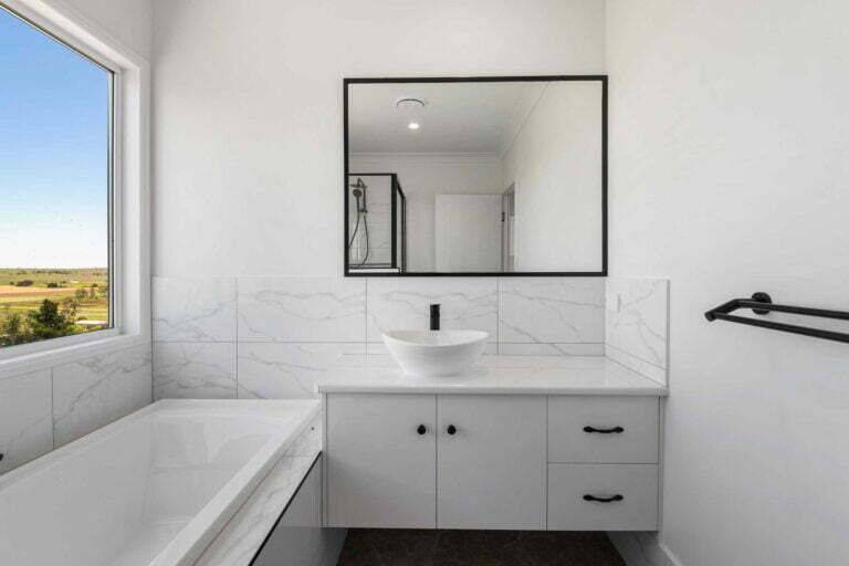 modern family bathroom with a single vanity unit, bath tub, large window overlooking acreage. Tiles are light and marbled, fixtures are black.